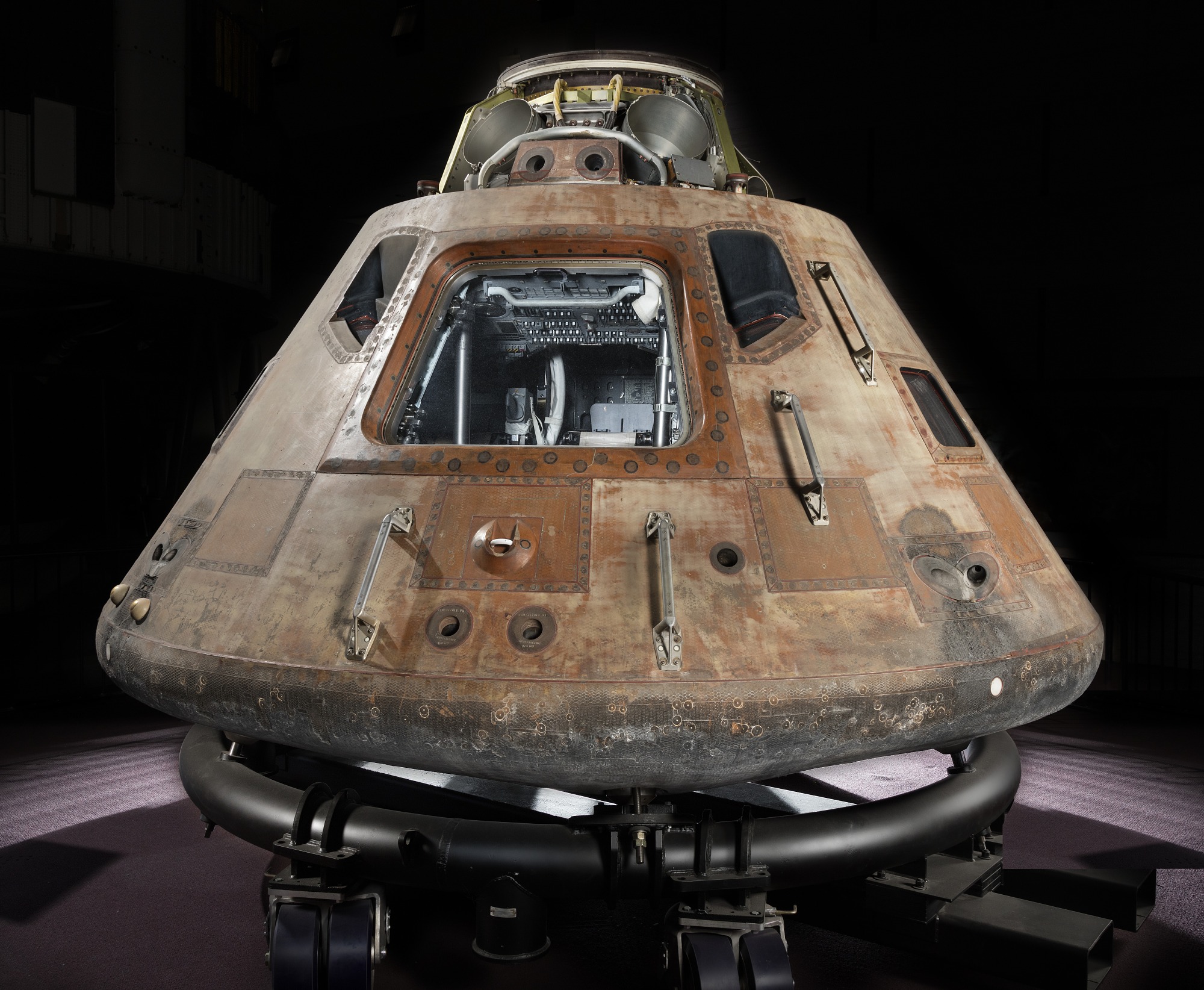 Conical-shaped spacecraft, photographed in a studio, constructed from numerous metal panels riveted together, now a rusted brown colour. Closest to the viewer is a rectangular hatch, with several small ‘side’ windows.